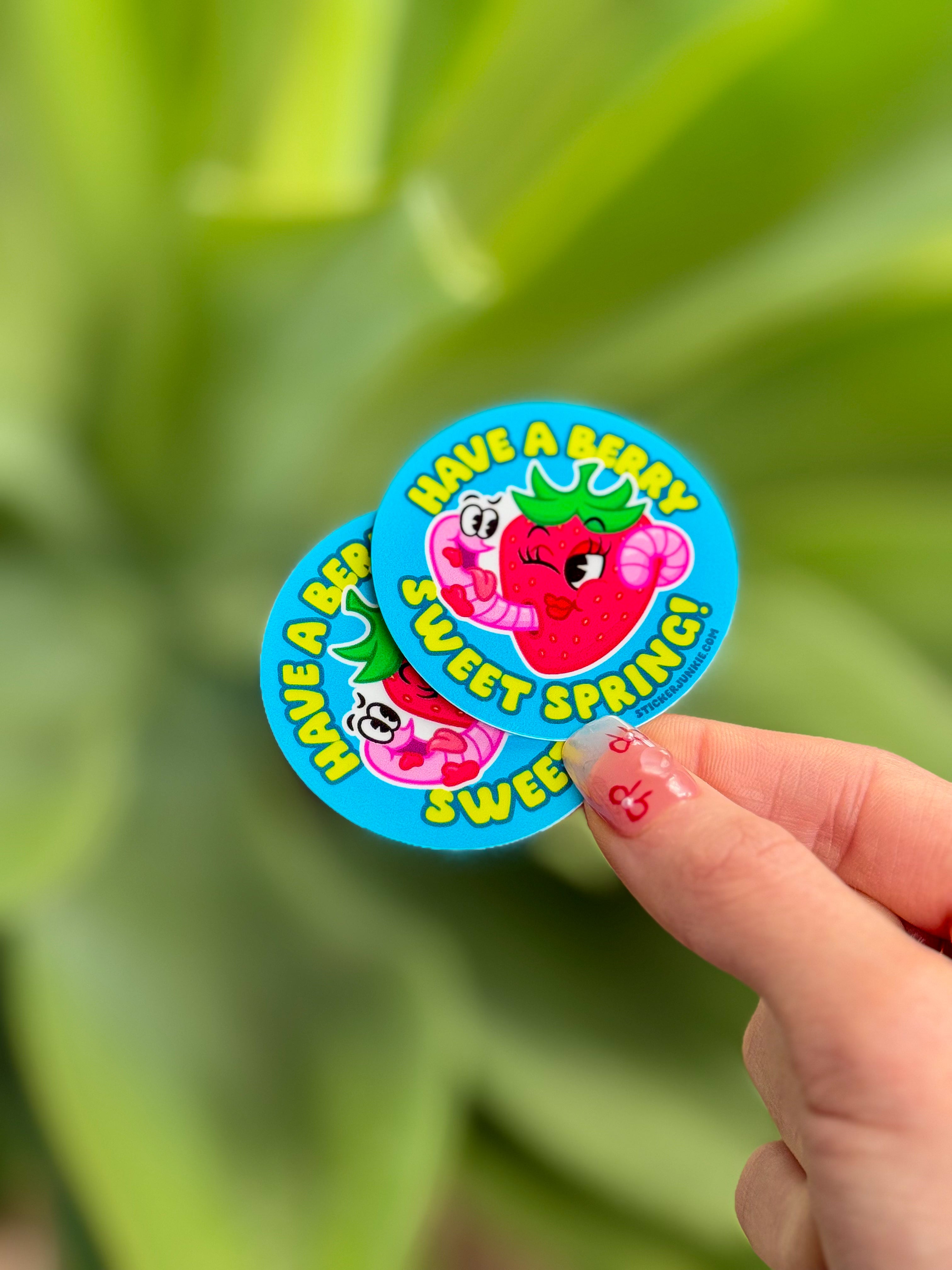 A hand holds two colorful, round stickers with cartoon strawberries and the words "Have a berry sweet spring!" against a blurred green leaf background.