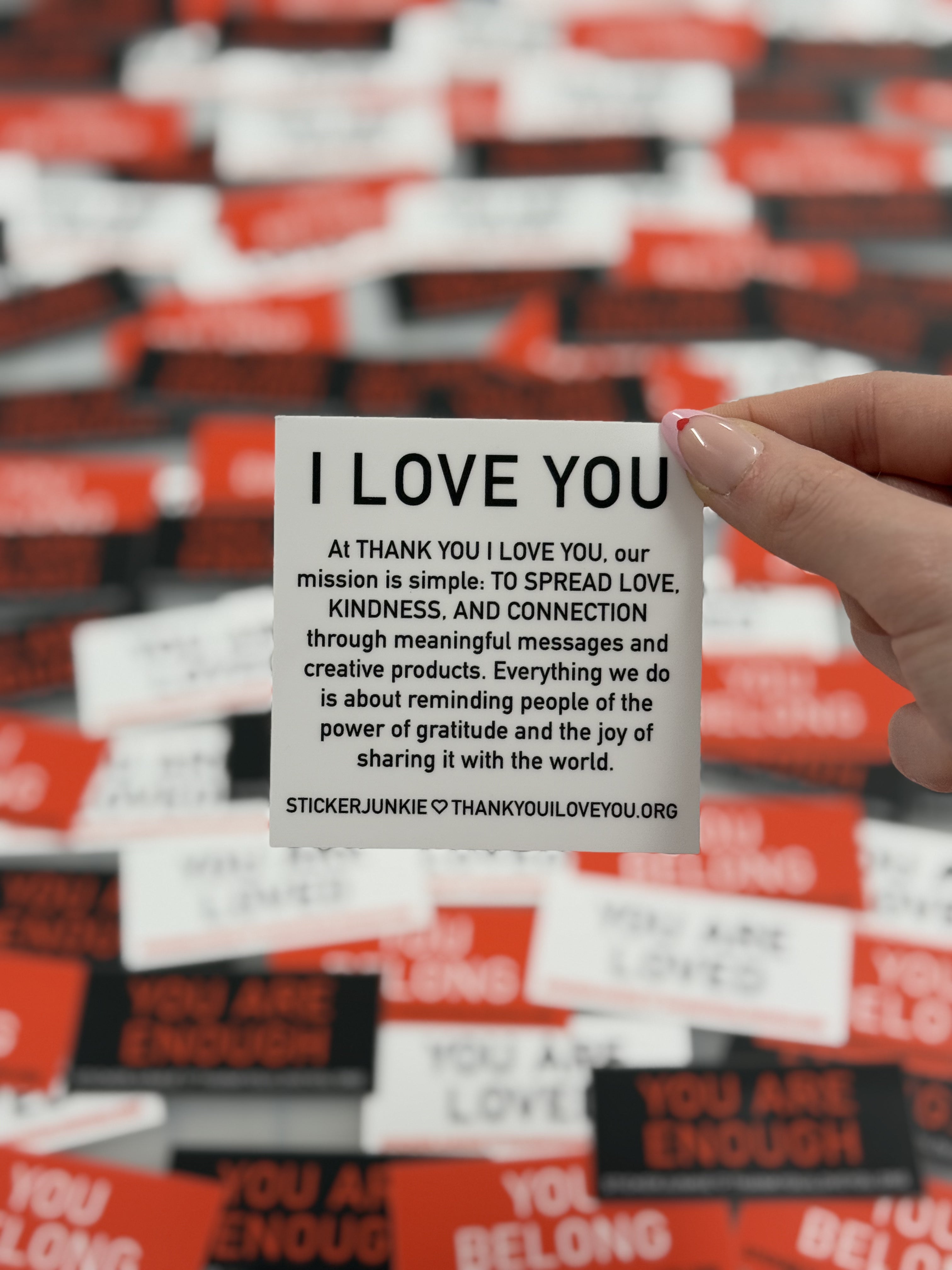 A hand holds a card with the message "I LOVE YOU" and a mission statement about spreading love and kindness. Background shows blurred inspirational stickers.