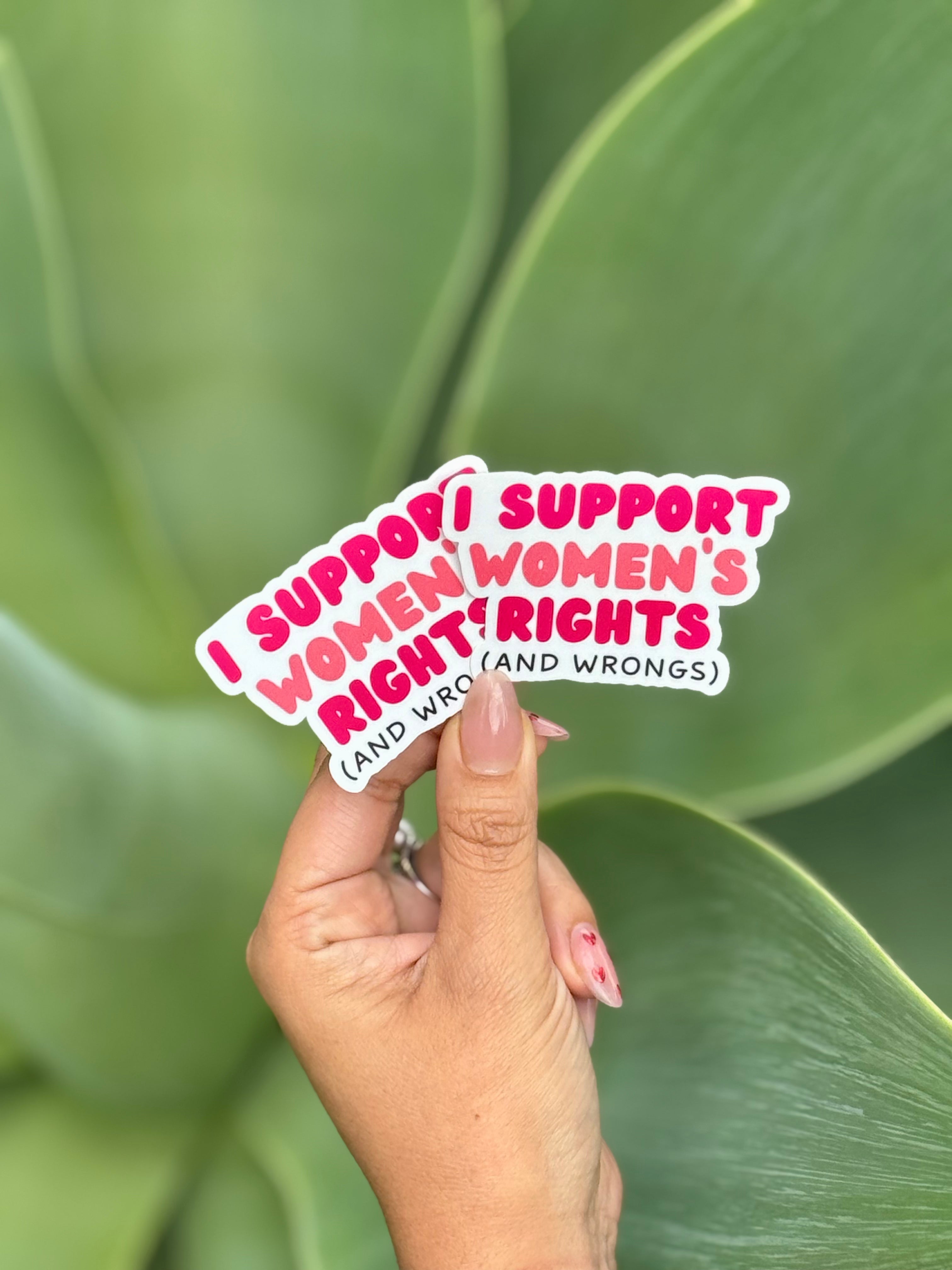 A hand with painted nails holds a custom die cut sticker that reads, "I support women's rights (and wrongs)" against a blurred green leaf background.
