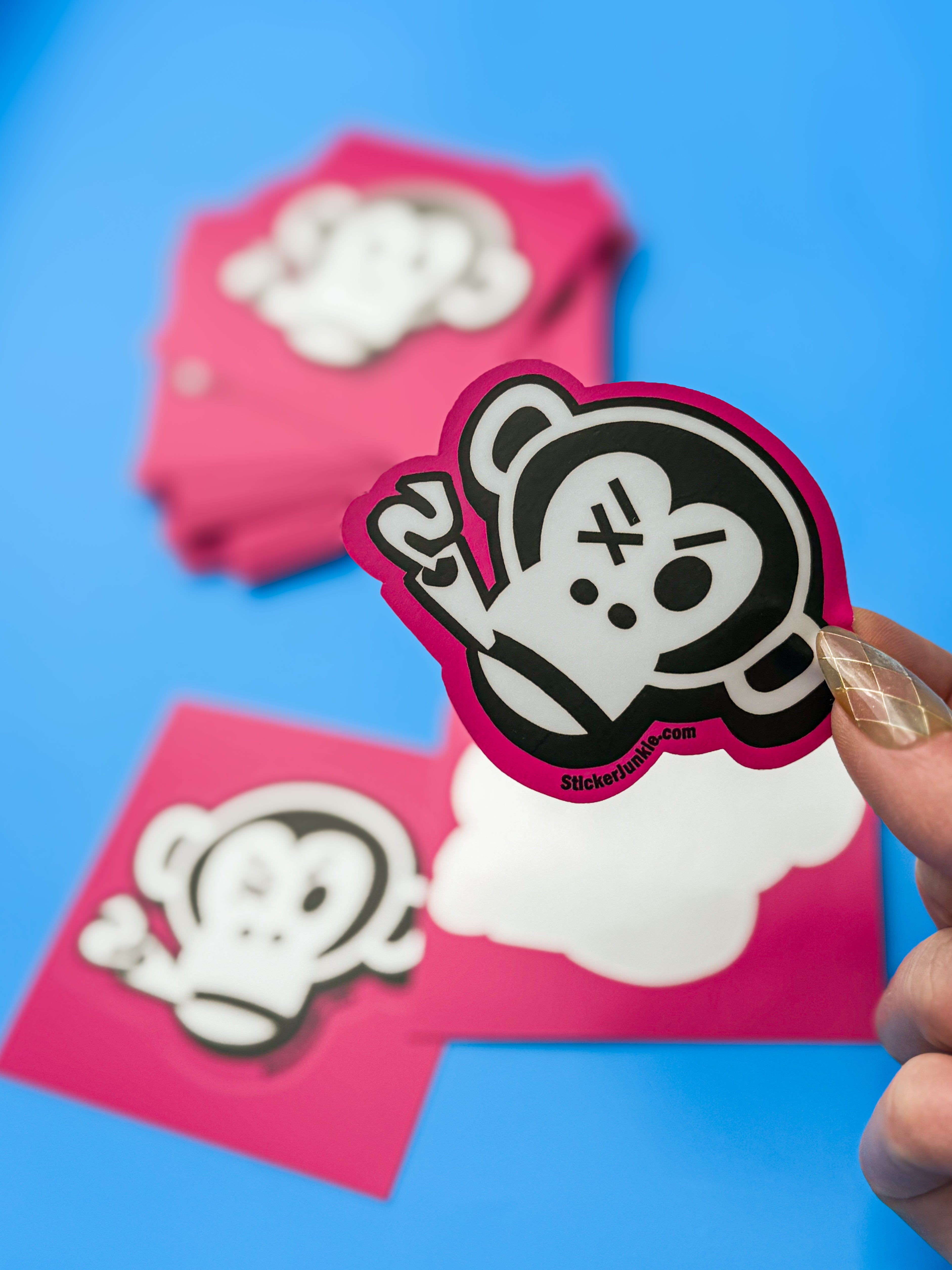 A hand holds a white and black monkey sticker with a playful expression on a pink background. More identical stickers lie slightly blurred below on a blue surface.