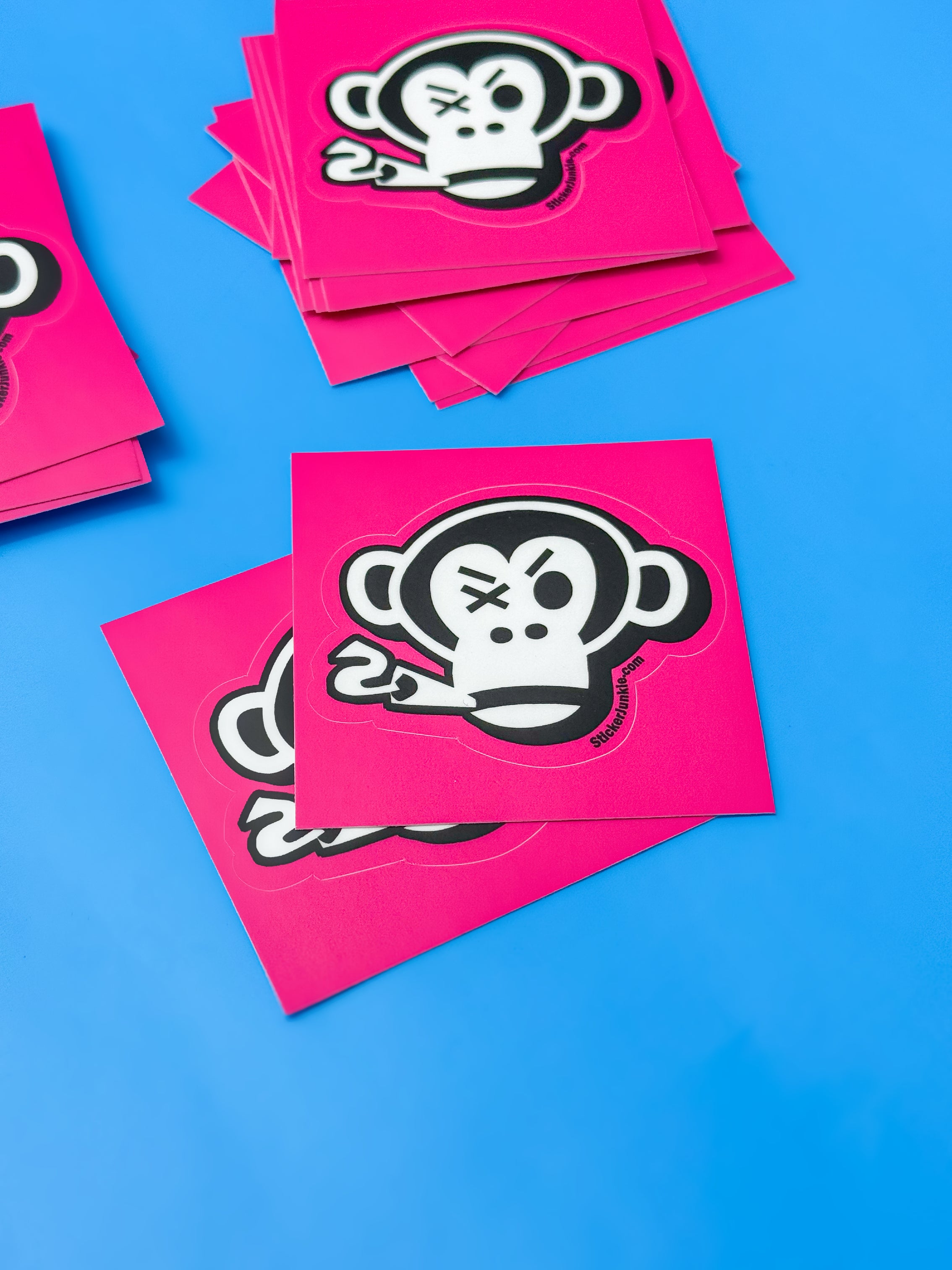 Bright pink stickers featuring a stylized white monkey with an eye patch, on a vivid blue background. The design is bold and playful.