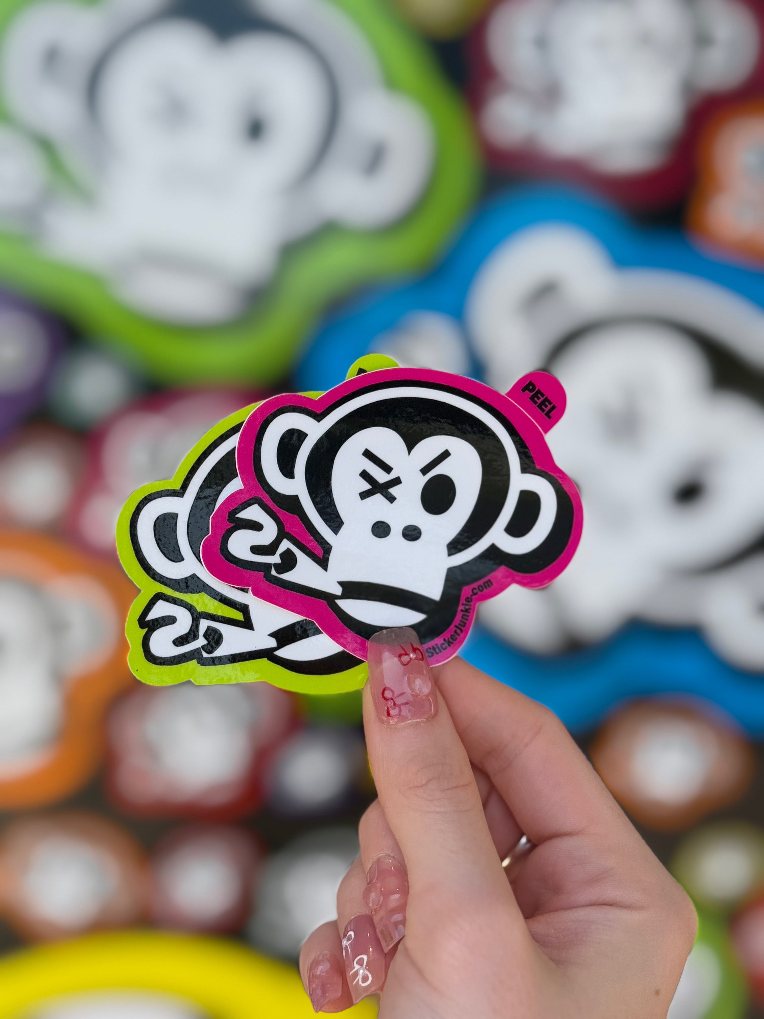 A hand with manicured nails holds colorful cartoon monkey peel tab stickers. The playful design has a vibrant background featuring other monkey images.