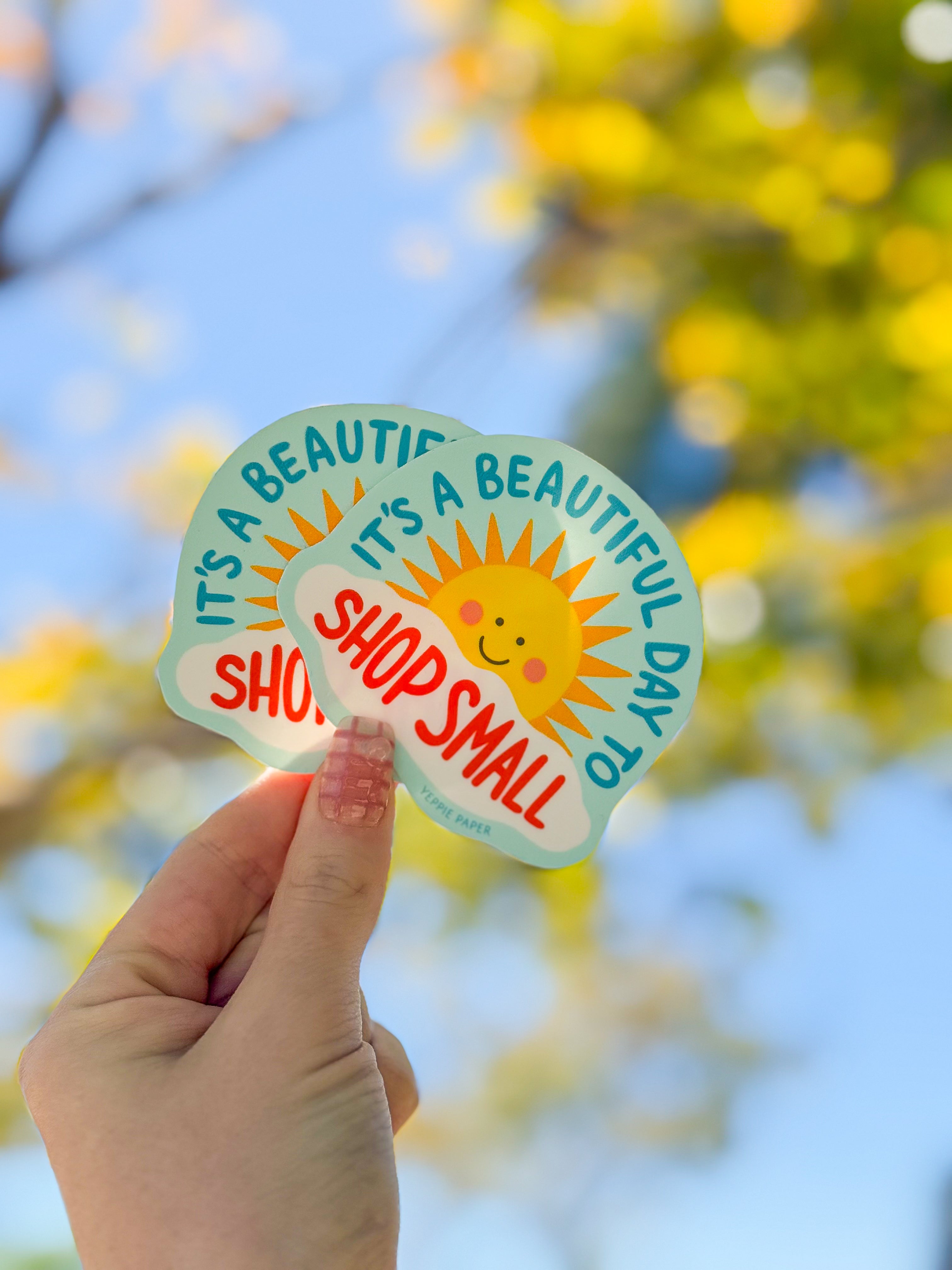A hand holds a cheerful sticker featuring a smiling sun and the words "It's a beautiful day to shop small," set against a sunny background with blurred foliage.