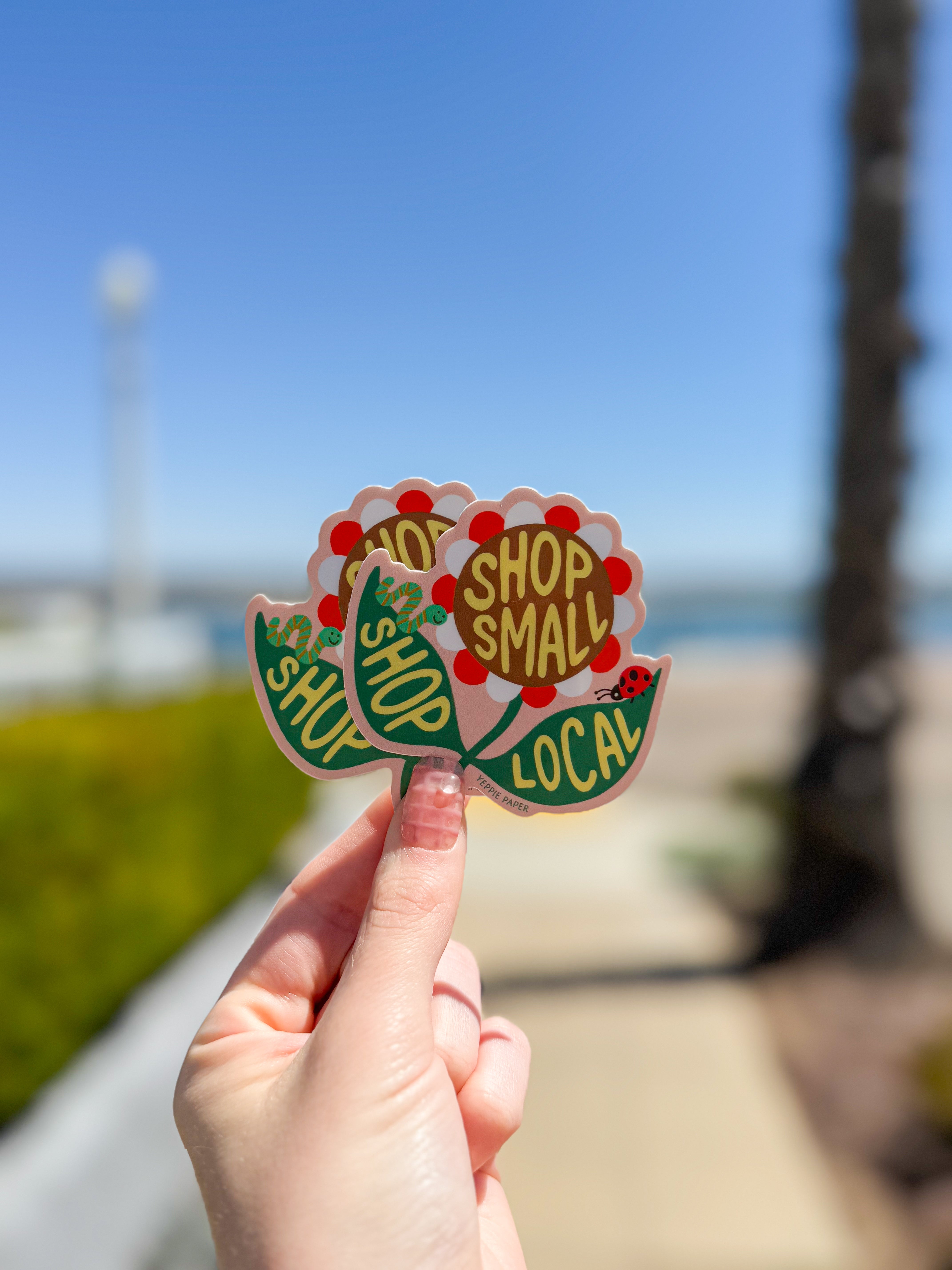 A hand holds two stickers with the text "Shop Small" and "Shop Local" in a floral design. Background shows a sunny, blurred street setting.
