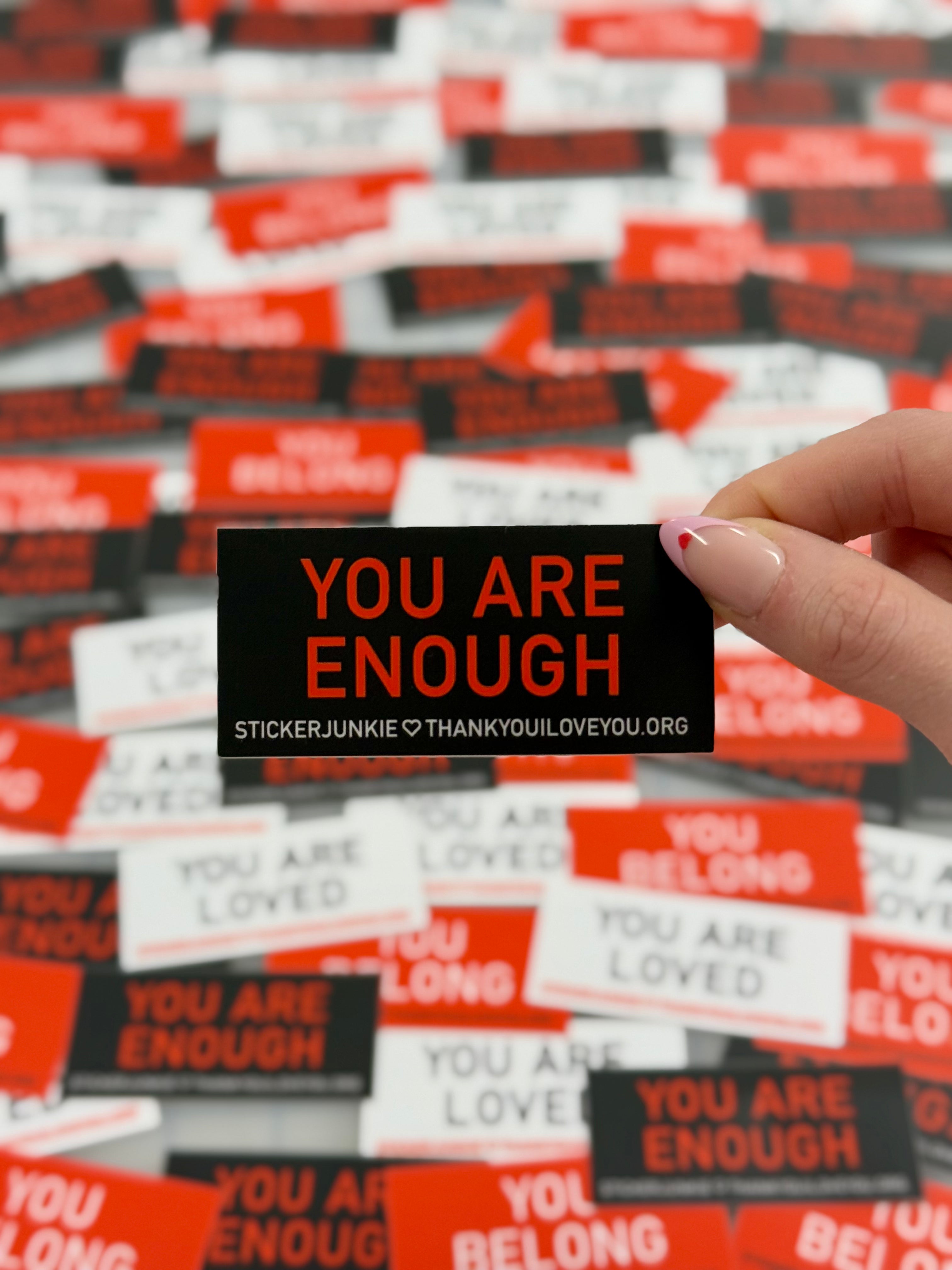 A hand holds a black sticker with "YOU ARE ENOUGH" in bold orange text. Similar motivational stickers are scattered in the blurred background.