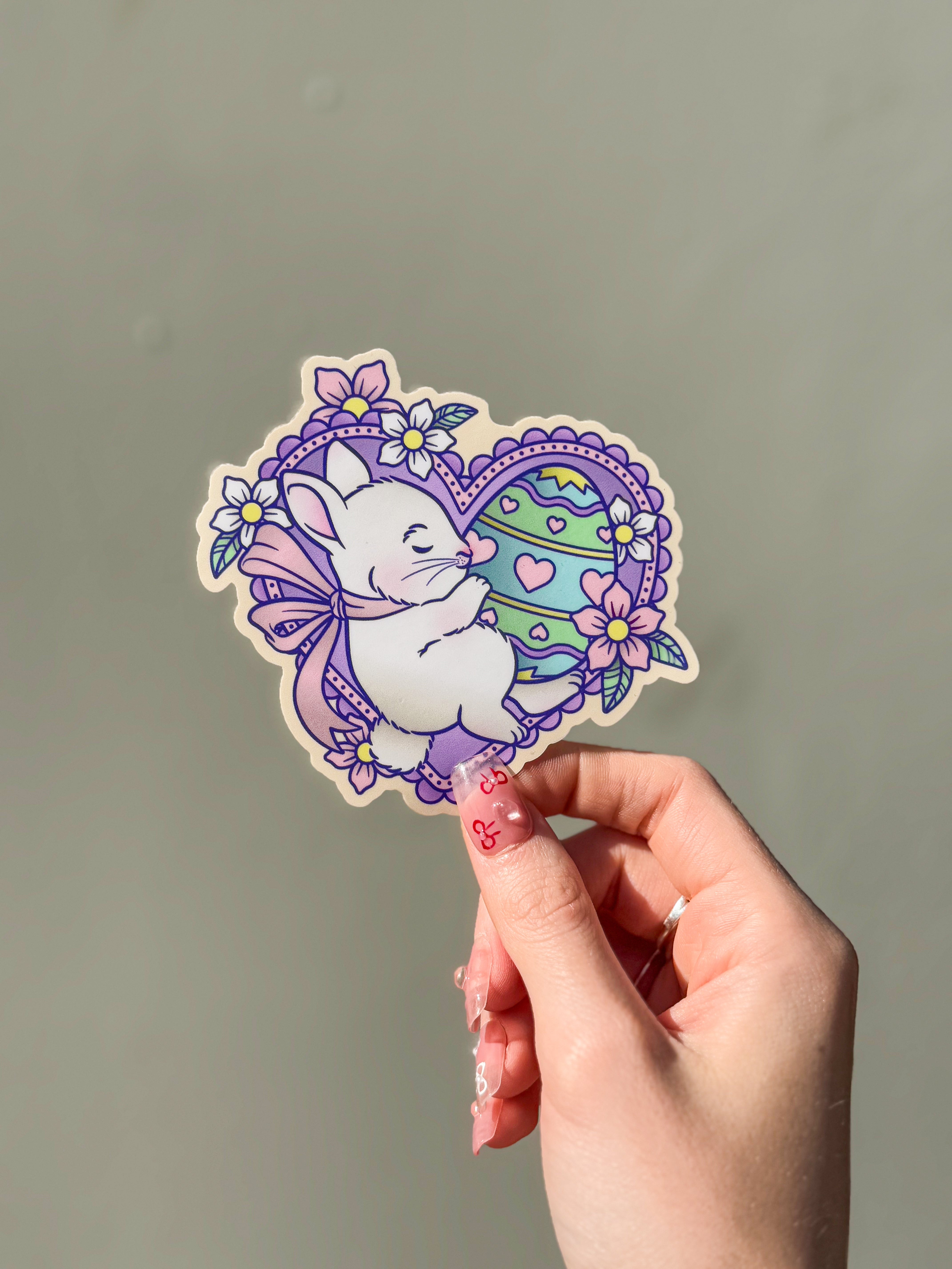 Example of a custom die cut sticker showing a white bunny hugging a decorated Easter egg, framed by a purple lace heart with floral details.