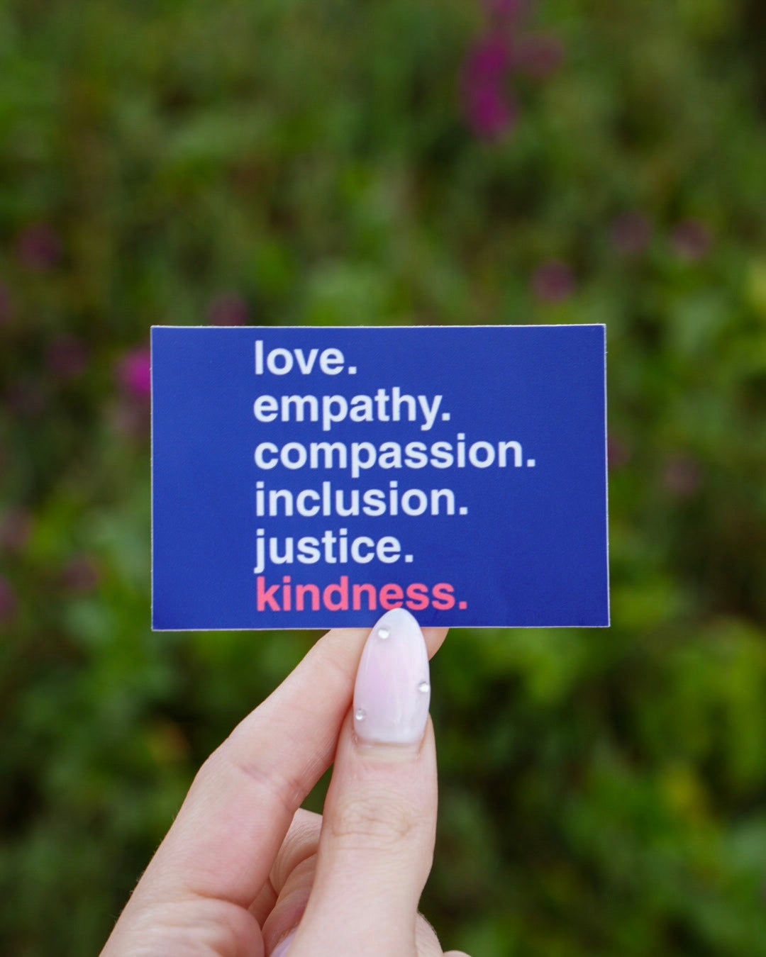 A hand holds a blue card with the words “love. empathy. compassion. inclusion. justice. kindness.” The background is blurred greenery.