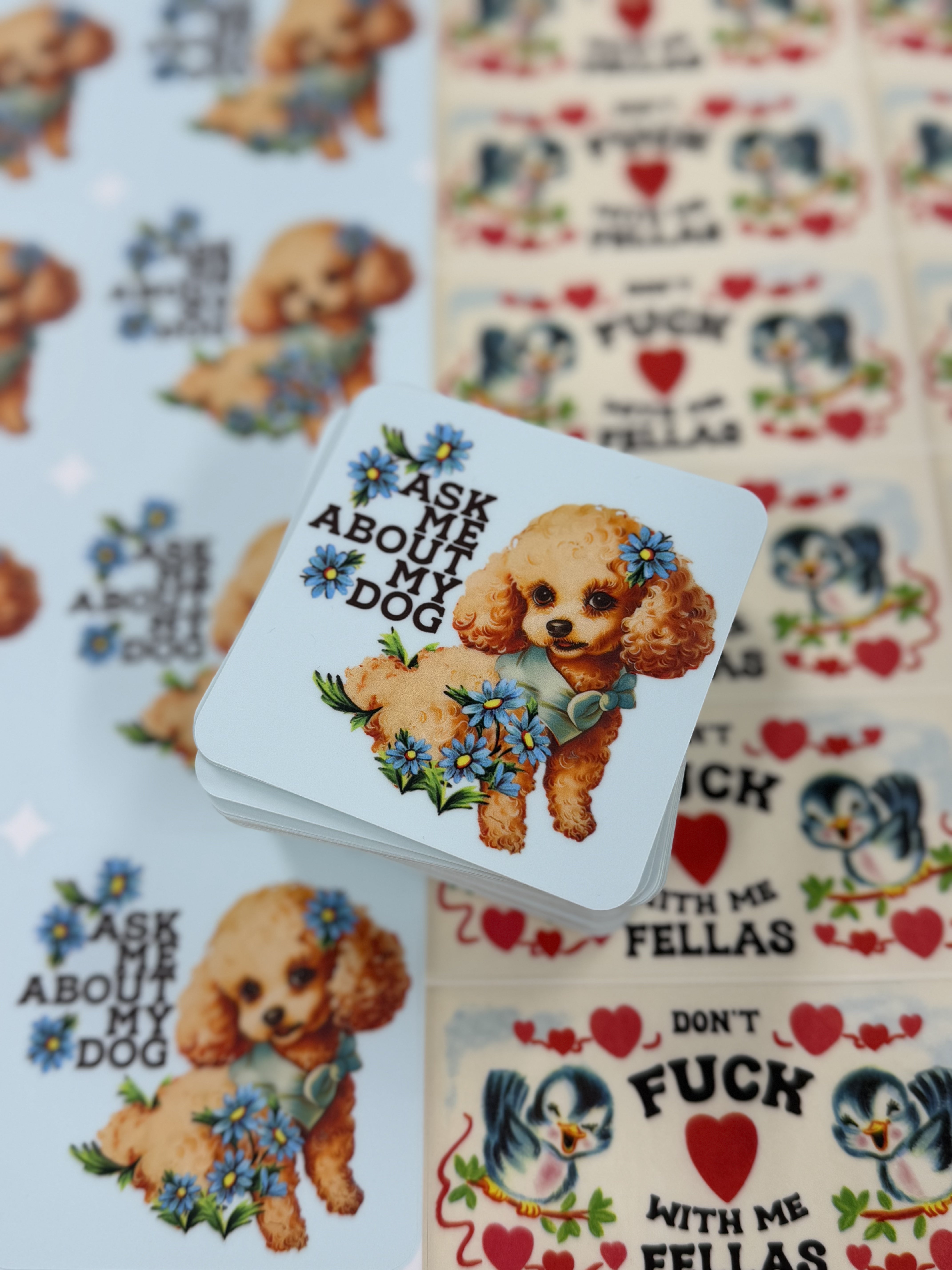 A stack of stickers featuring a cute poodle with blue flowers and text "Ask Me About My Dog" overlap bird-themed stickers that say "Don't F**k With Me Fellas," set on a pastel background.