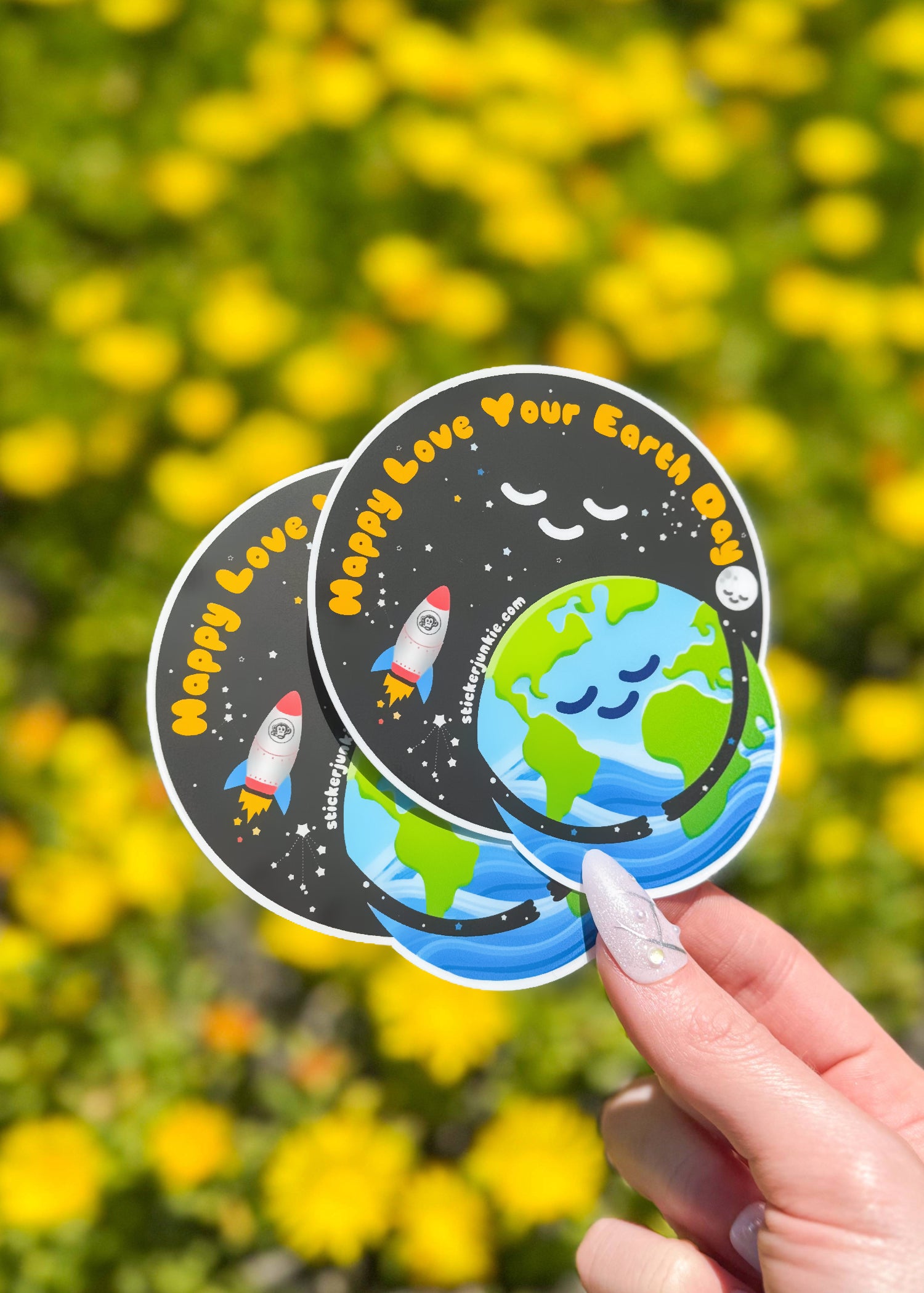 A hand holding two circular stickers that say "Happy Love Your Earth Day," featuring a smiling Earth and a rocket, with yellow flowers blurred in the background.