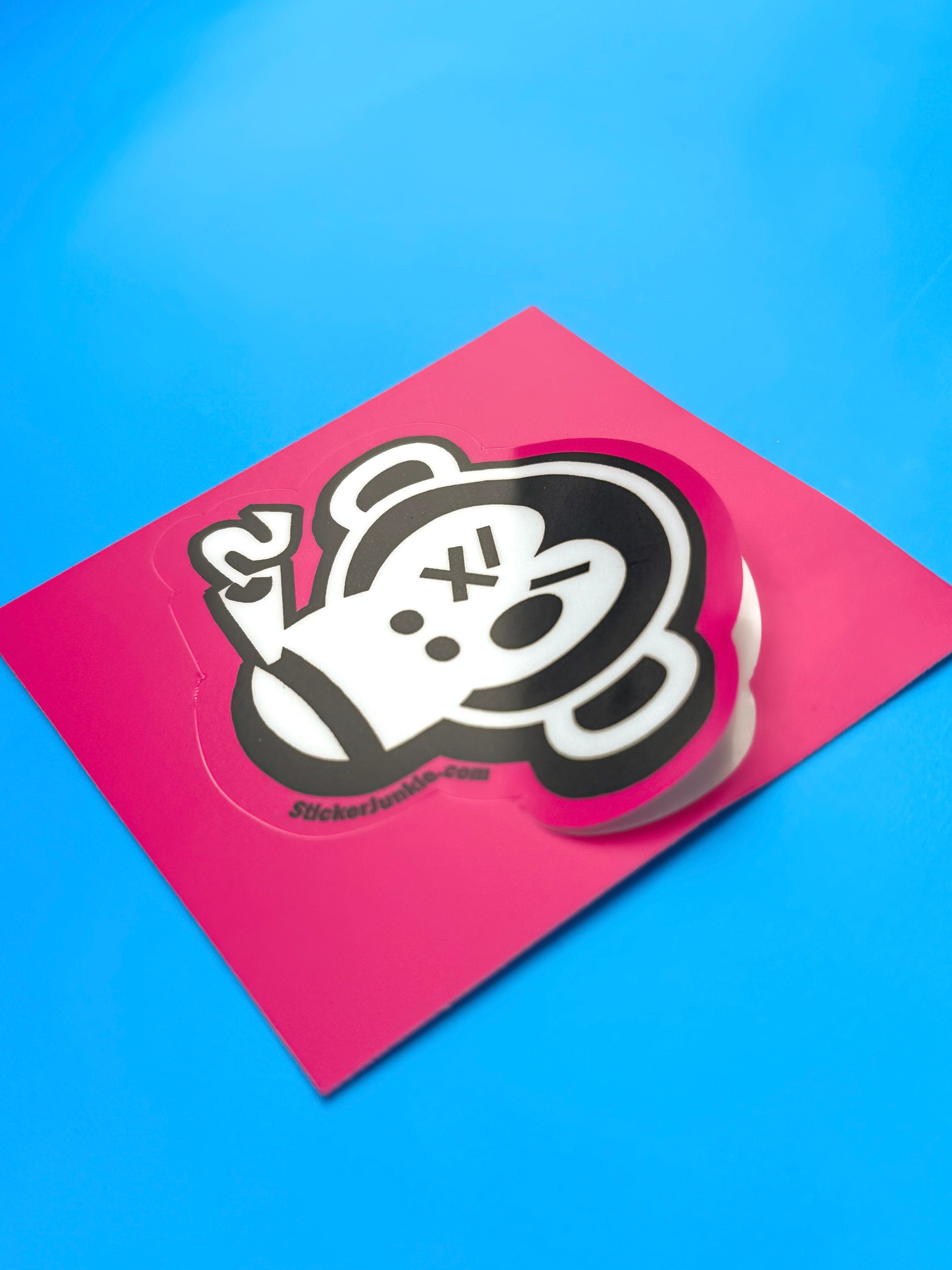 A playful black and white monkey kiss cut sticker with a quirky expression, surrounded by a bright pink background on a blue surface.