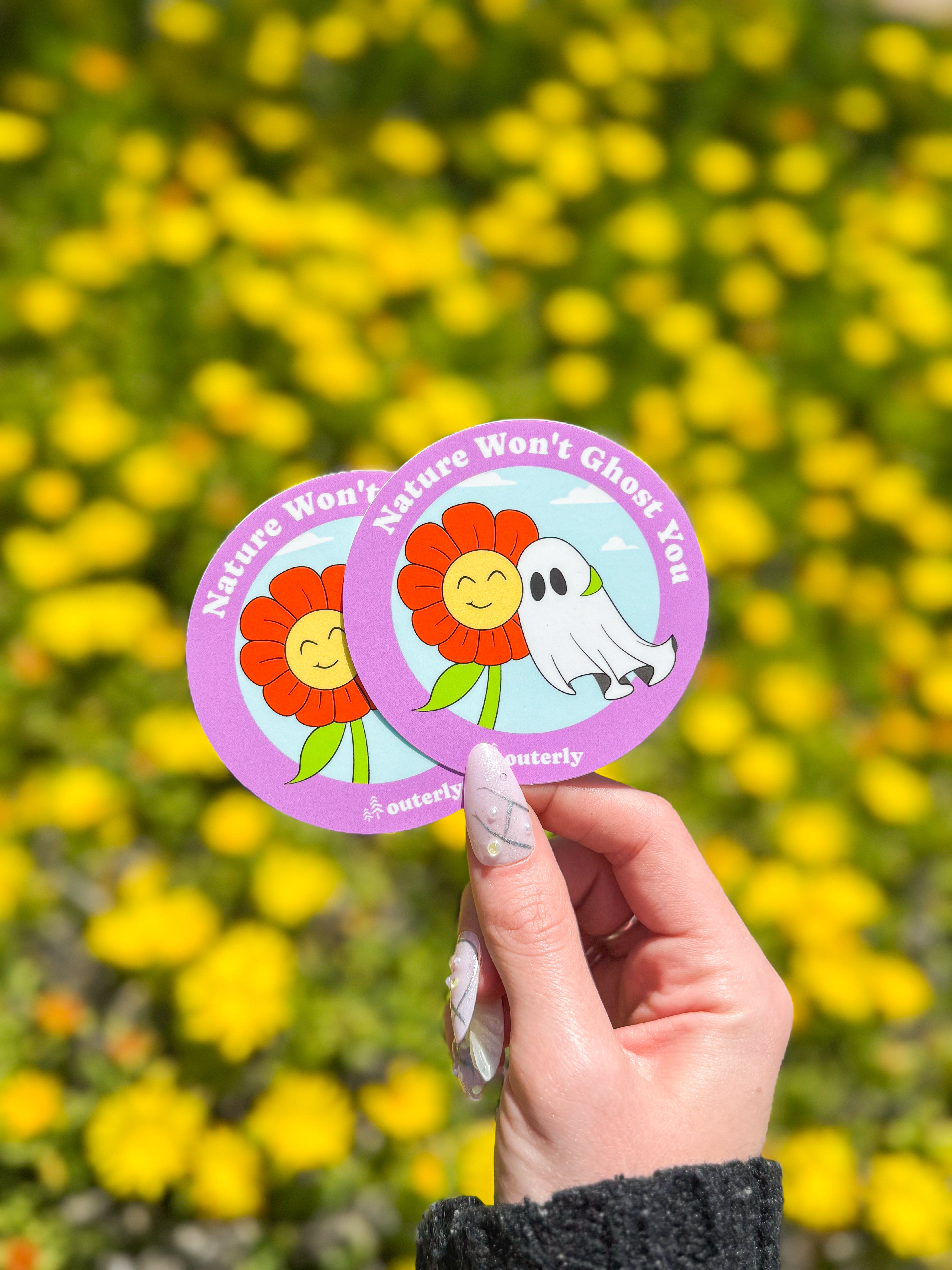 A hand with manicured nails holds two round stickers against a backdrop of yellow flowers. Stickers feature a smiling flower and ghost with text "Nature Won't Ghost You."