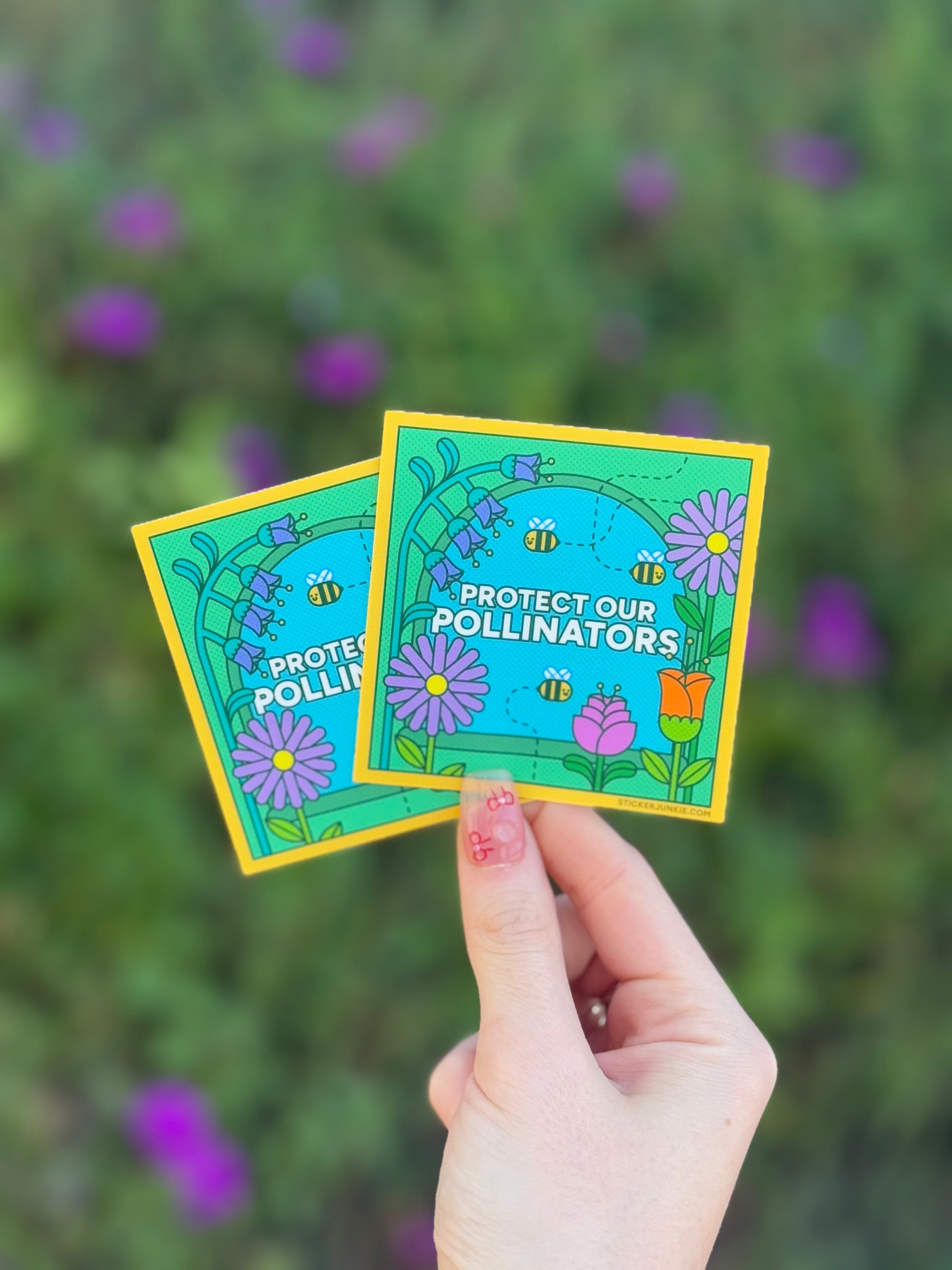 A hand holds two colorful stickers with flowers and bees, titled "Protect Our Pollinators," against a blurred green background with purple flowers.