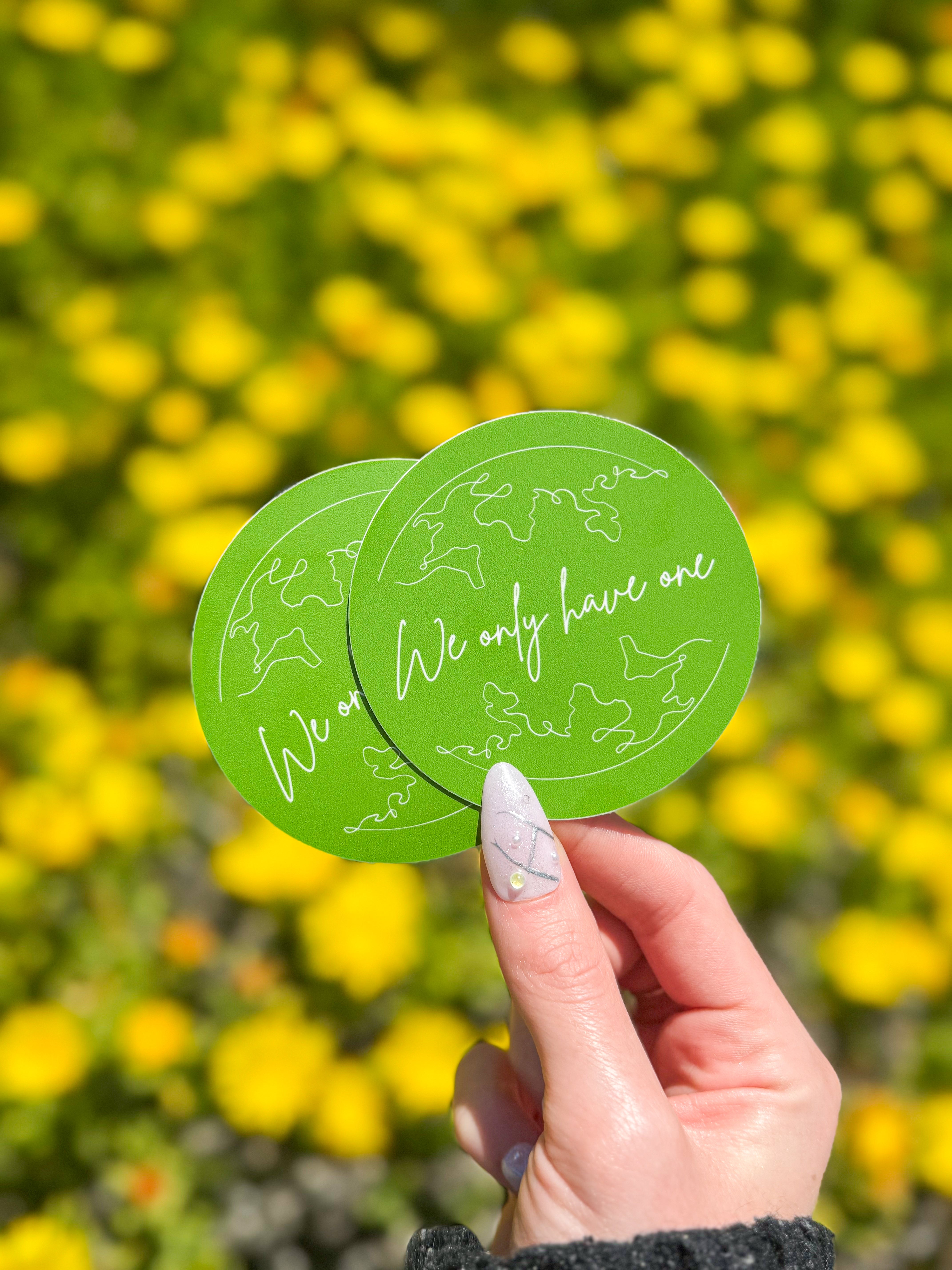 A hand with manicured nails holds two green circular stickers with white outlines of continents and the text "We only have one," against a blurred background of yellow flowers.