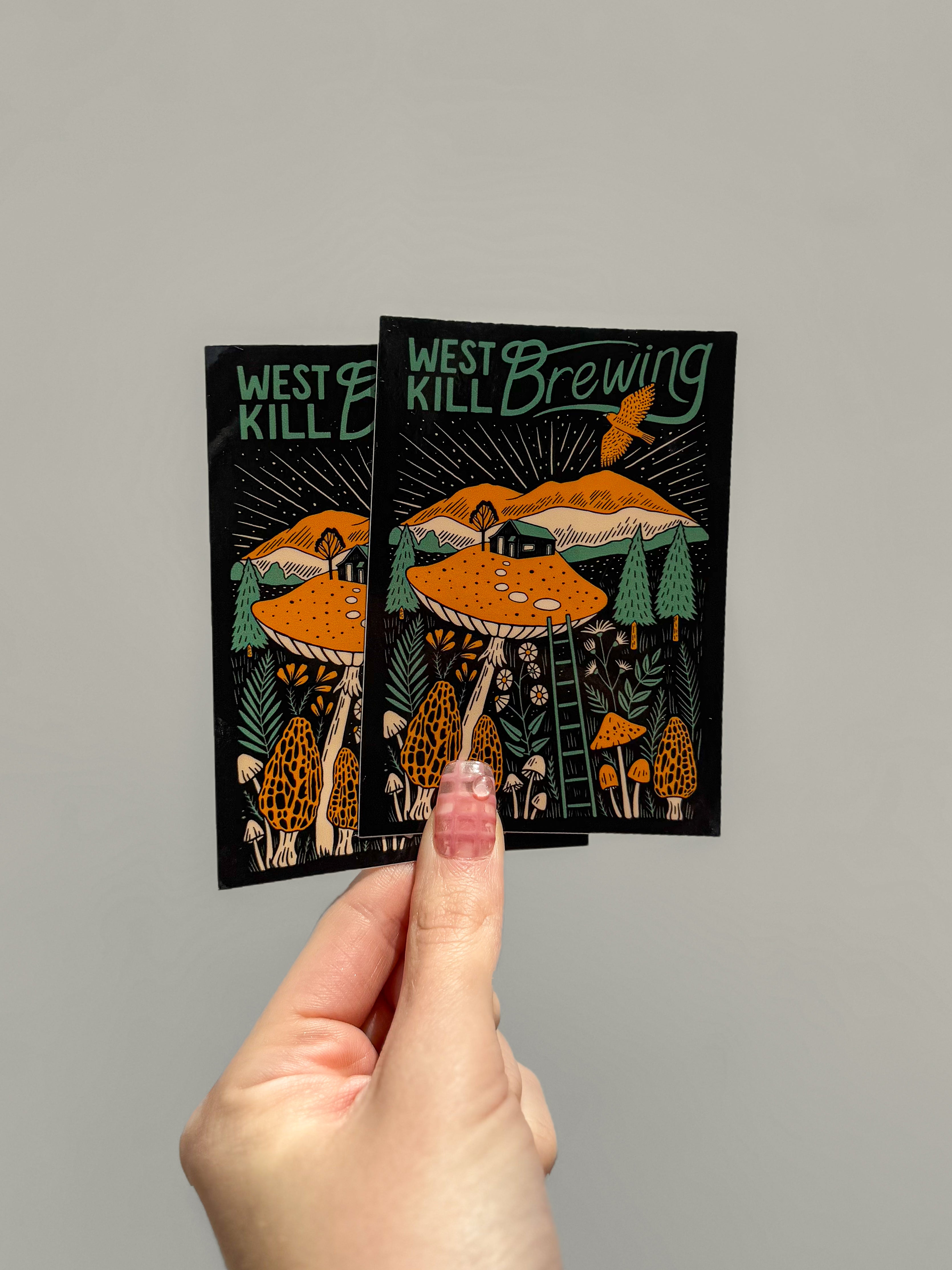 A hand holds two identical rectangle stickers with vibrant designs of mushrooms, trees, and a mountain. The text reads "West Kill Brewing" with an artistic style.