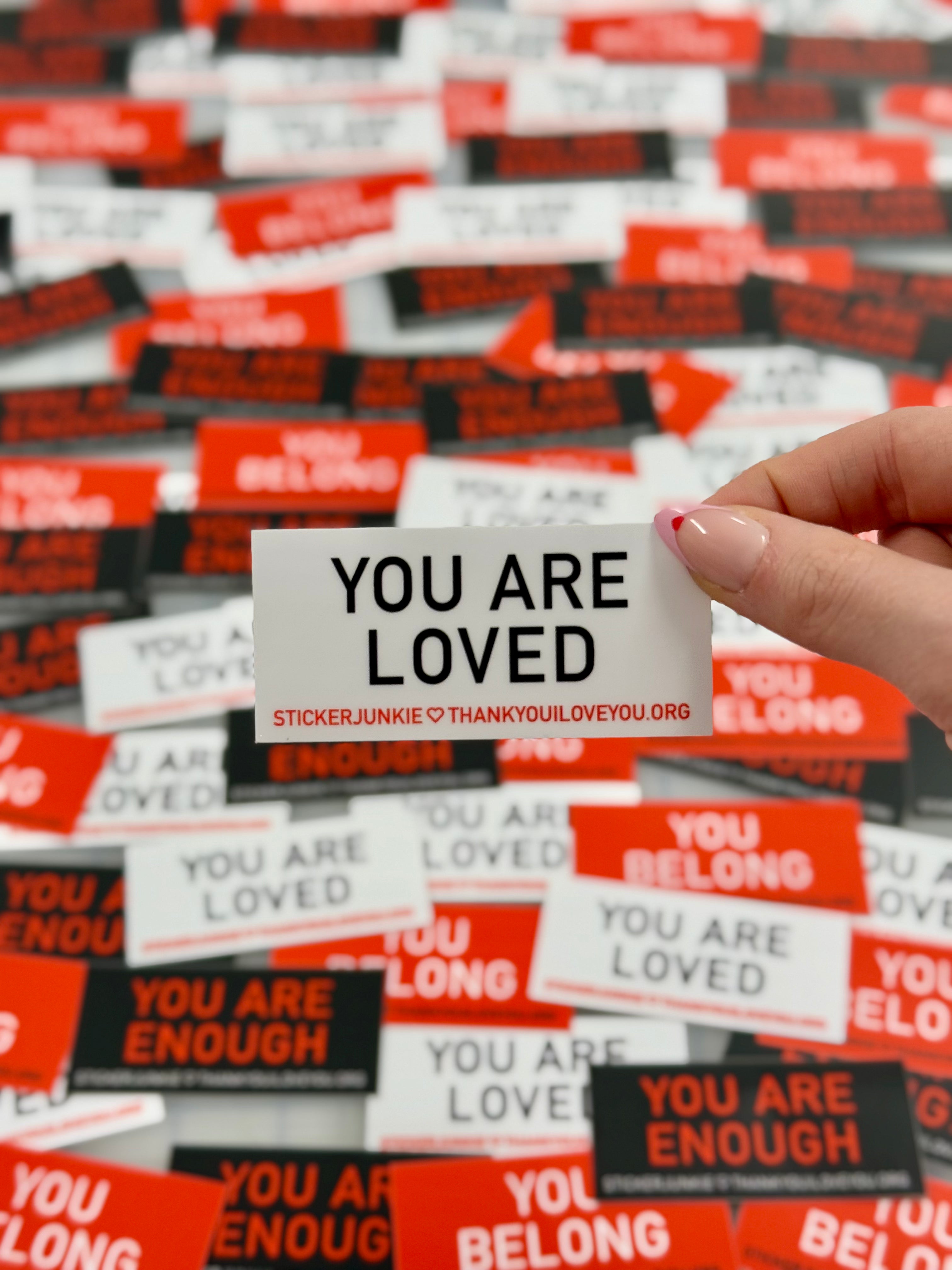 A hand holding a sticker with "You Are Loved" text. Background filled with similar motivational stickers in red, black, and white, creating a positive, uplifting mood.